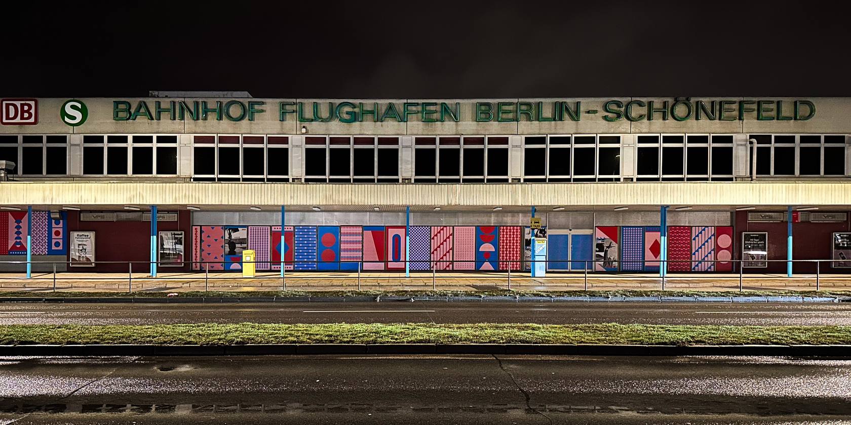 Schönefeld station’s entrance building at night: a low building with the historic lettering “Bahnhof Flughafen Berlin-Schönefeld” and colourful patterned artwork on the ground-floor façade, with a road in front.