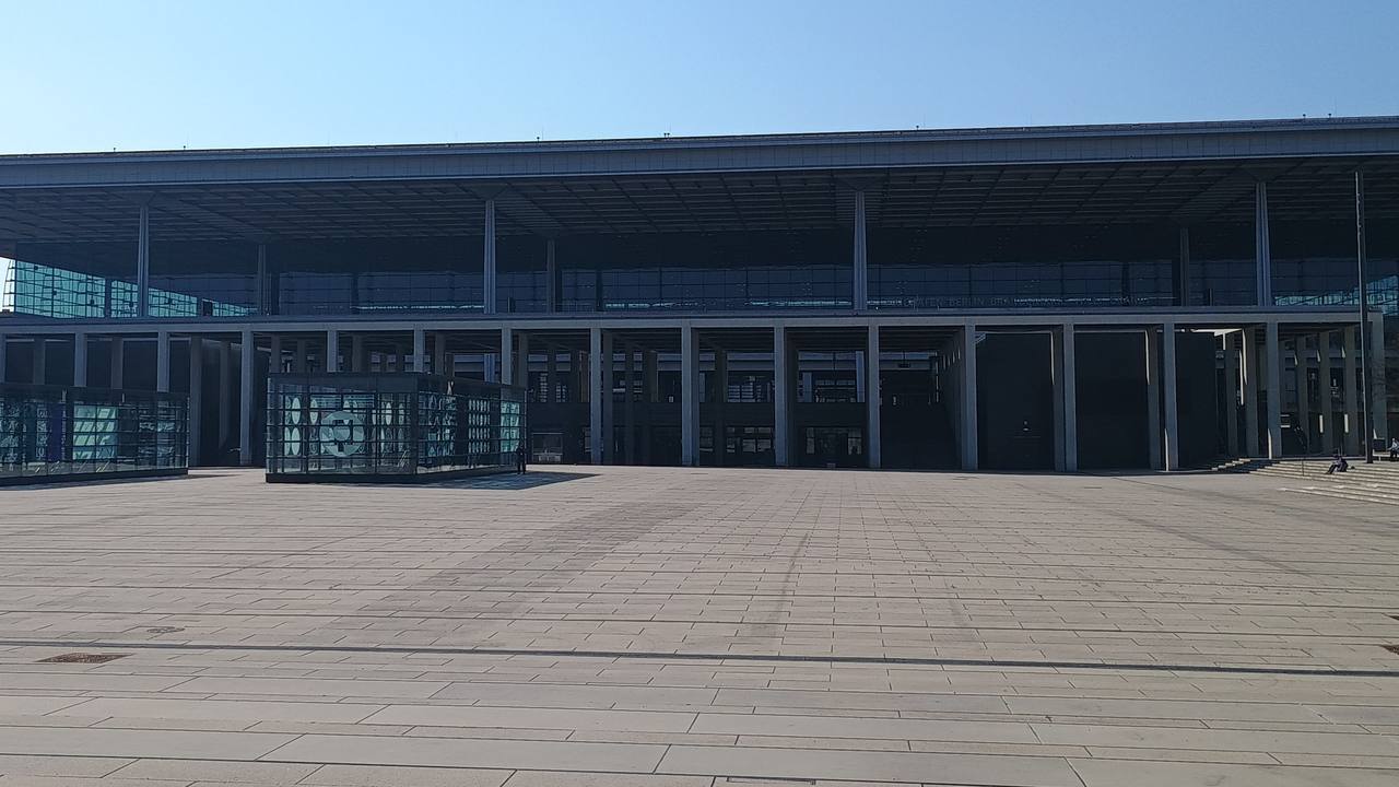 Terminal 1 at BER Airport: a long, low glass façade under a wide projecting roof, with the broad paved Willy-Brandt-Platz in front under a blue sky.